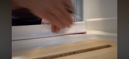 A close-up of a hand applying or smoothing a white adhesive strip with a thin red edge onto a white surface, with a wooden board in the foreground.